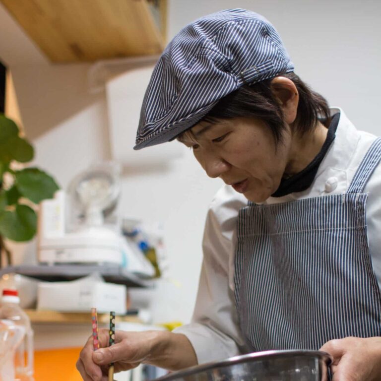 Kazue.O。コック帽と縞のエプロン姿で、厨房で手元の鍋に向かっている